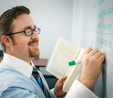 man writing on board with green marker while holding book in other hand