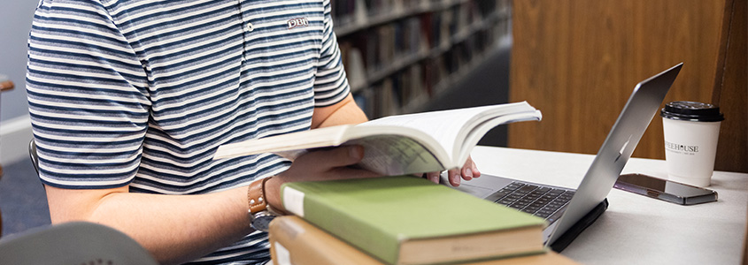 DBU student reading a book and computer in library